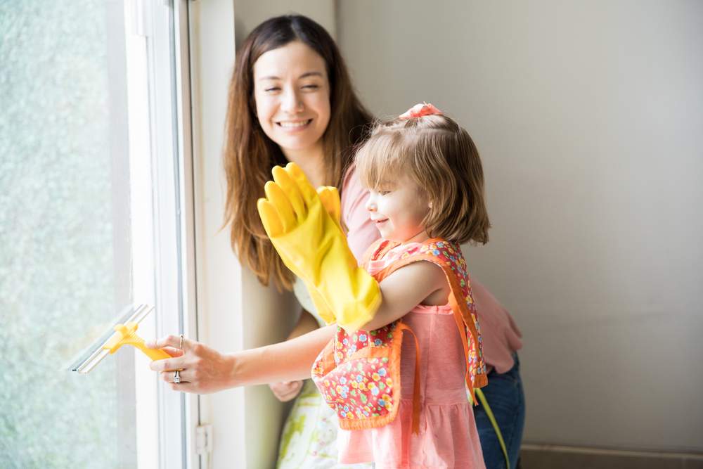 Cute adorable girl claps using gloves while her pretty mother cleans the window from inside the house