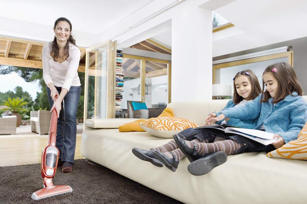 Young mum using a vacum cleaner wthile her two twin daughters look at a book in the living room.