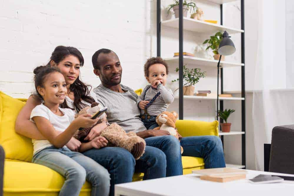 little girl holding remote while sitting on the sofa watching tv with her family.