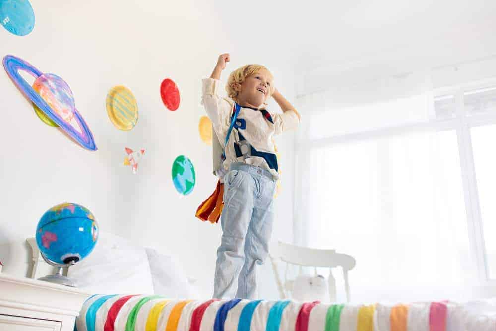 Kids play astronaut. Little boy in space costume jumping on bed with rocket. Solar system and planet room decoration. Creative child, future profession.