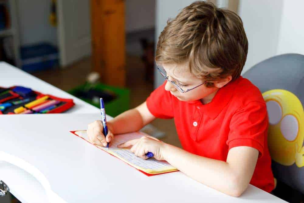 Cute little kid boy with glasses at home making homework, writing letters with colorful pens. Little child doing exercise, indoors. Elementary school and education, imagine fantasy concept.