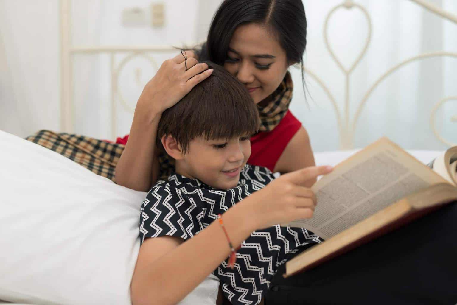 Mom teach son reading a book is on the bed at home.