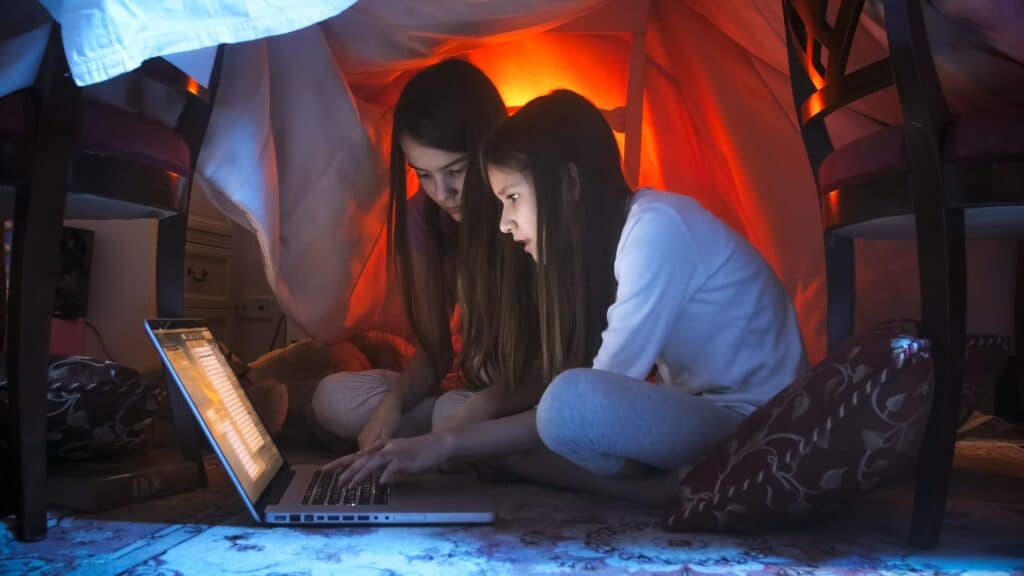 Two sisters in pajamas sitting in tent at bedroom with laptop watching clean movies