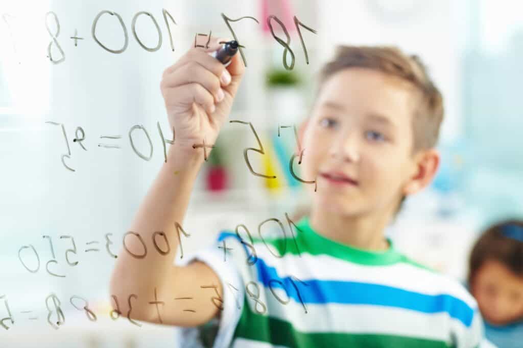 Boy looking at a clear board and doing math with a marker.