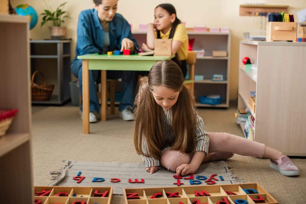Lady sitting with a girl at a desk while another learns with letters on the floor.