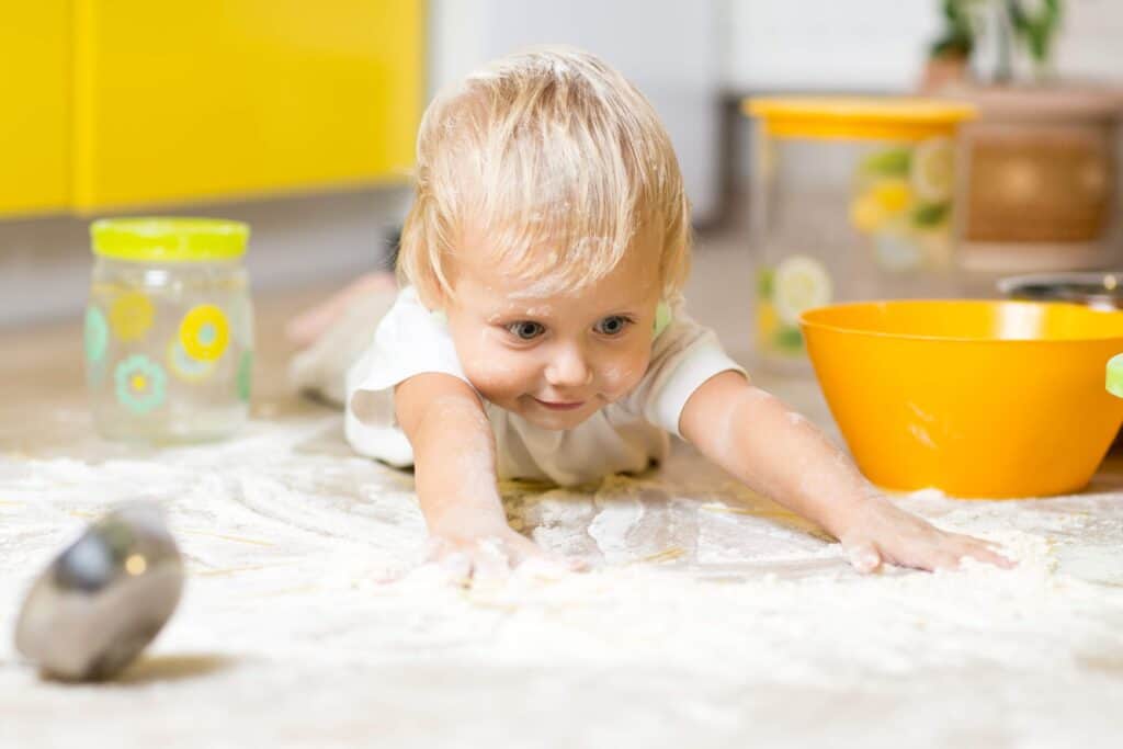 Little boy child laying on very messy kitchen floor. Toddler covered in white baking flour.