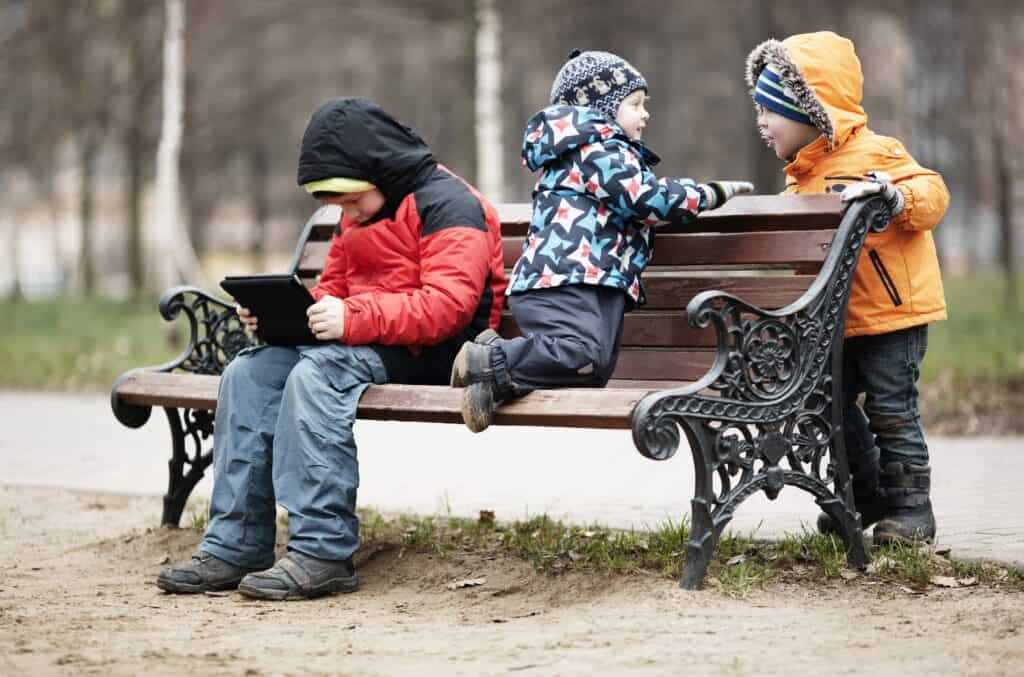 Three young boys playing on a park bench in winter wrapped up warmly against the cold weather with one youngster reading a tablet as the two younger children romp around