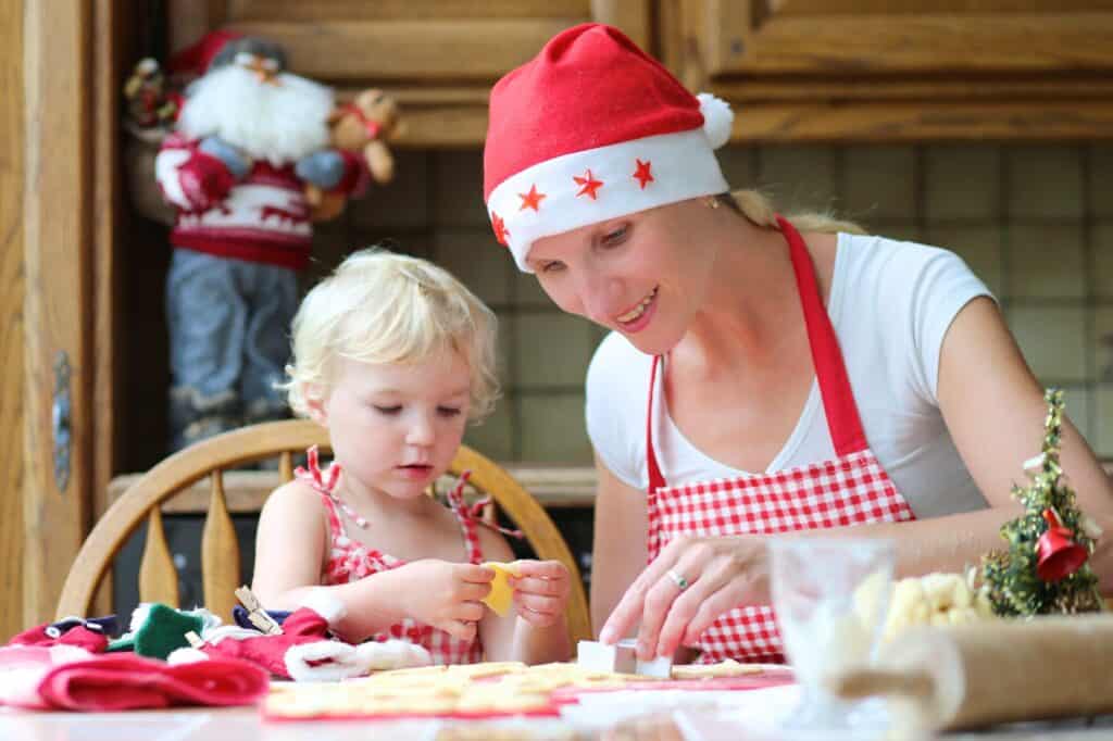 Happy mother, young woman in red apron and santa hat baking christmas cookies together with her little daughter, cute blonde toddler girl, sitting at the table in classic traditional wooden kitchen